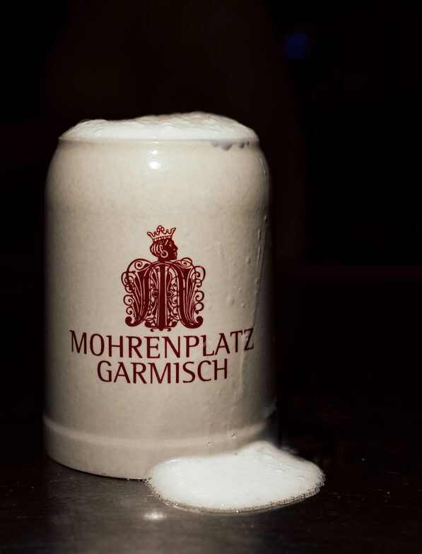 Steinkrug mit übergelaufenem Bierschaum und rotem Schriftzug „Mohrenplatz Garmisch“ vor dunklem Hintergrund