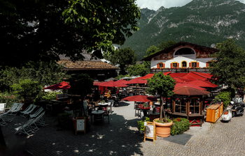 Schattiger Biergarten mit roten Schirmen, Holzbar und Liegestühlen vor bemaltem Wirtshaus und bewaldetem Berg