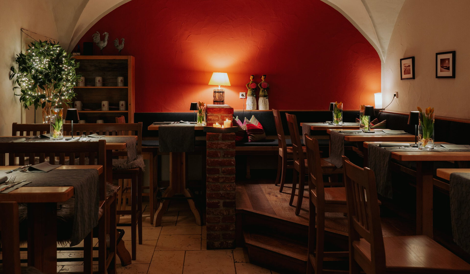 Abendlich beleuchteter Gewölberaum mit roter Stirnwand, Holztischen, Tulpen und Kerzenleuchter an der Decke