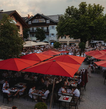 Blick von oben auf den vollen Biergarten unter roten Schirmen und einen Platz voller weiß gekleideter Festgäste